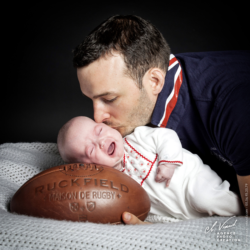 Photo studio d'un bébé allongé sur un ballon de rugby pendant que son père l'embrasse